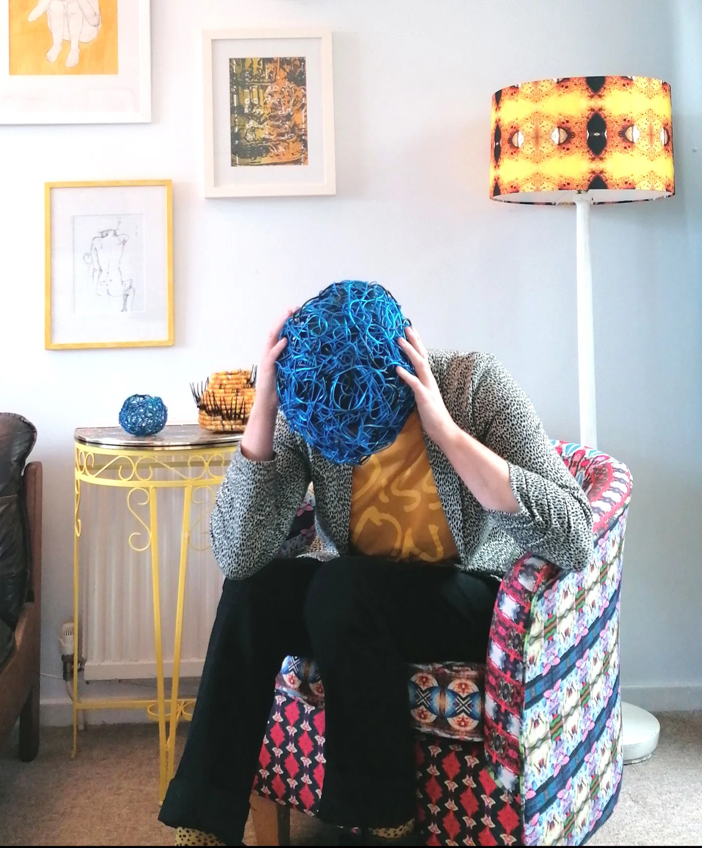 Artist wears a blue wire basket over her head and rests head in hands.